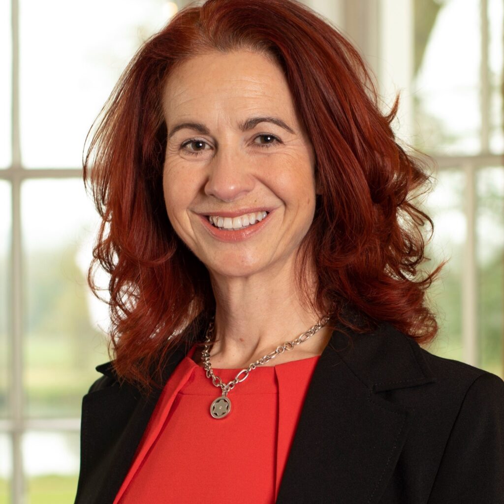 Smiling lady with red hair, wearing a red top and black jacket.