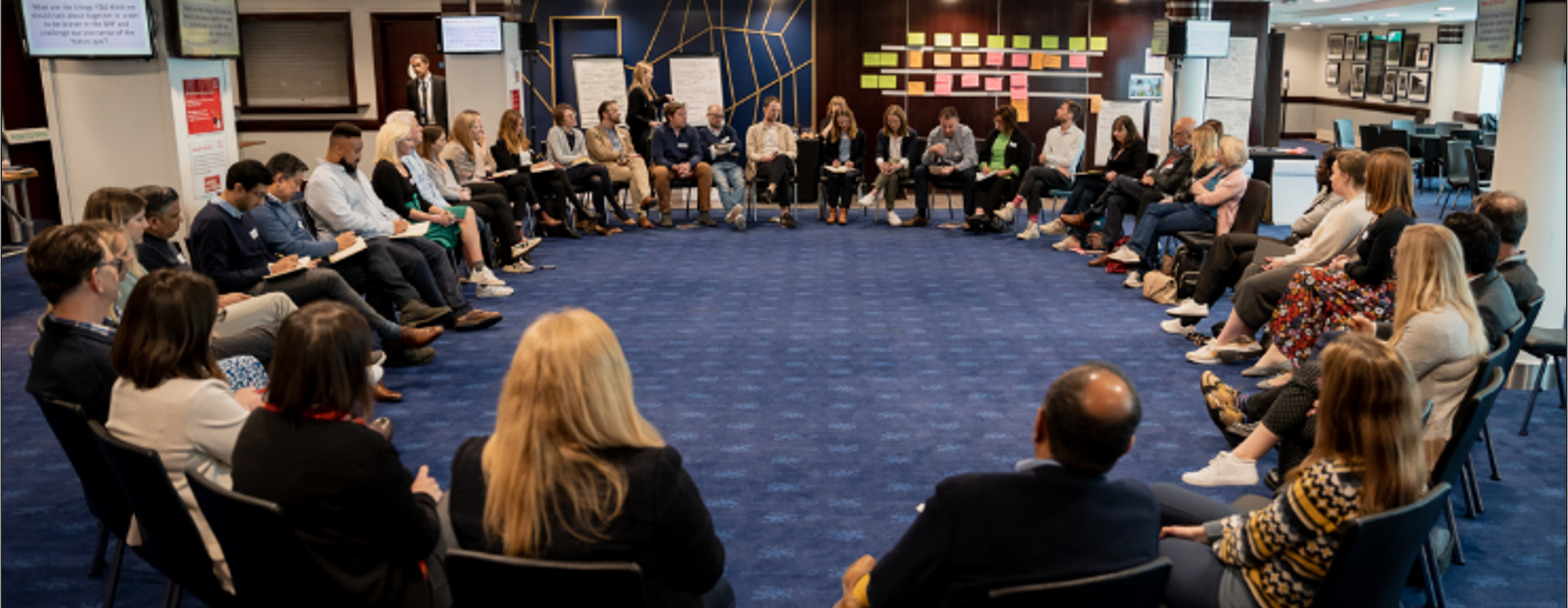 a large group of people sat in a a circle of chairs in a large event room